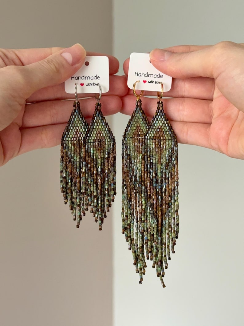 May include: Two pairs of long, beaded fringe earrings featuring a geometric pattern in shades of green, brown, and blue. One pair has silver-toned hoops, and the other has gold-toned hoops. Each pair is attached to a white card labeled Handmade with love.
