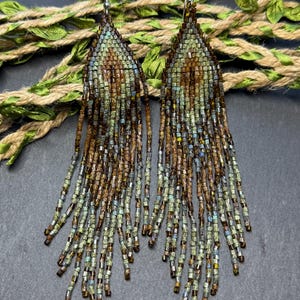 May include: A pair of long, teardrop-shaped fringe earrings crafted from small glass beads in shades of sage green, bronze, and brown. The earrings feature a woven triangular top section that transitions into multiple dangling beaded strands.