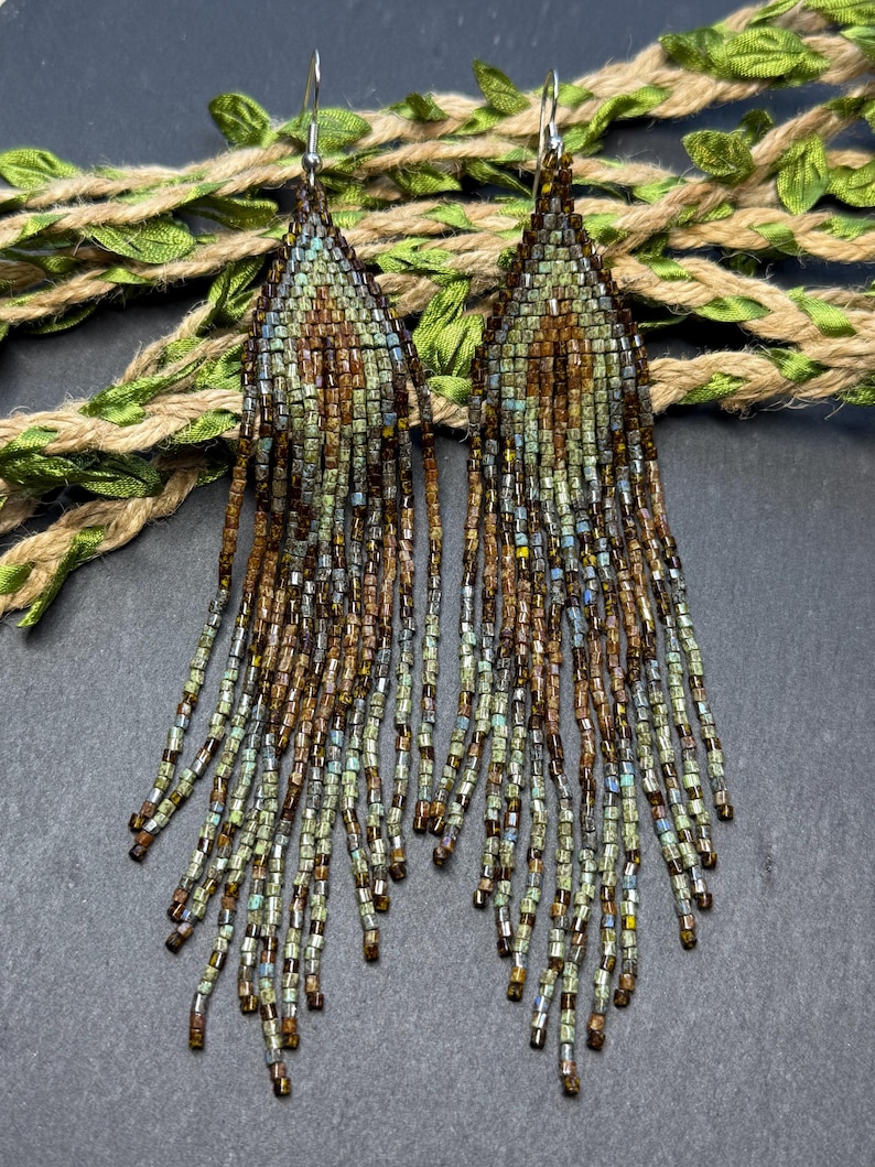 May include: A pair of long, teardrop-shaped fringe earrings crafted from small glass beads in shades of sage green, bronze, and brown. The earrings feature a woven triangular top section that transitions into multiple dangling beaded strands.