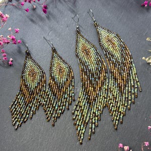 May include: Two pairs of long, tapered fringe earrings crafted from small glass beads in shades of bronze, copper, and sage green. The earrings feature a woven pattern that transitions into loose, dangling strands, suspended from silver-toned hooks.