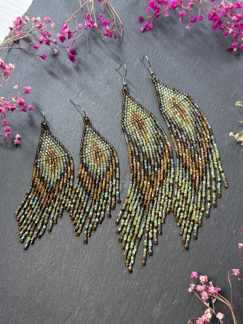 May include: Two pairs of long, tapered fringe earrings crafted from small glass beads in shades of bronze, copper, and sage green. The earrings feature a woven pattern that transitions into loose, dangling strands, suspended from silver-toned hooks.