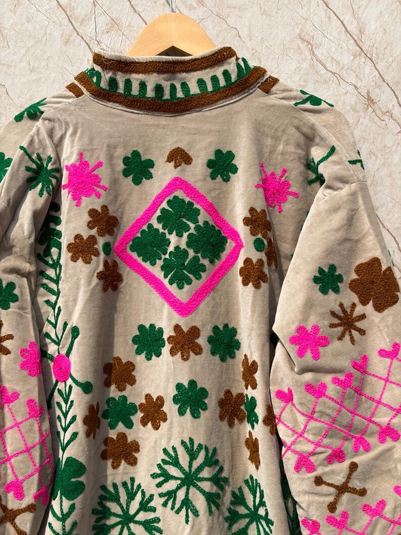 Puede incluir: T&uacute;nica de terciopelo beige con intrincados bordados en verde, marr&oacute;n y rosa intenso. El dise&ntilde;o presenta patrones florales y geom&eacute;tricos, incluyendo una forma de diamante con motivos verdes en forma de tr&eacute;bol. La t&uacute;nica tiene cuello alto y mangas largas.