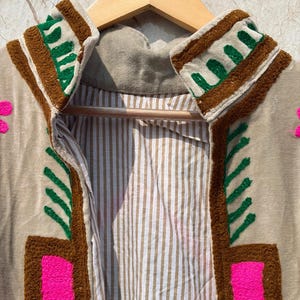 Puede incluir: Chaqueta beige bordada con detalles en marr&oacute;n, verde y rosa. La chaqueta tiene un forro interior a rayas y se muestra en una percha de madera. El bordado incluye patrones geom&eacute;tricos y florales.