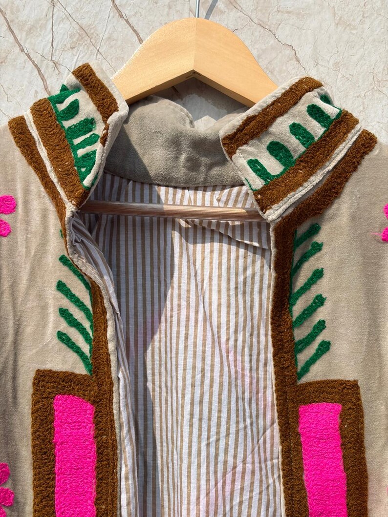 Puede incluir: Chaqueta beige bordada con detalles en marr&oacute;n, verde y rosa. La chaqueta tiene un forro interior a rayas y se muestra en una percha de madera. El bordado incluye patrones geom&eacute;tricos y florales.
