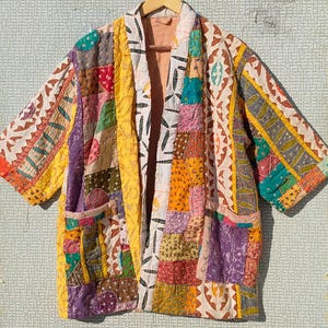 Può includere: Giacca patchwork colorata con maniche corte, appesa a una gruccia di legno. La giacca presenta una varietà di motivi e colori di tessuto, tra cui giallo, viola, arancione e verde. Ha due tasche.
