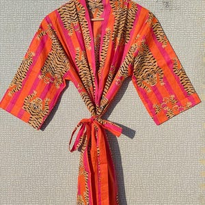 May include: A vibrant orange and pink robe with a tiger stripe pattern. The robe features a tie closure and is hanging on a wooden hanger. The design includes vertical stripes and tiger illustrations.
