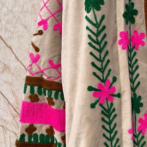 Puede incluir: Primer plano de una prenda beige bordada con detalles vibrantes. El dise&ntilde;o presenta patrones florales rosas y verdes y acentos geom&eacute;tricos. La manga tiene un patr&oacute;n de rayas marrones y rosas. La prenda est&aacute; hecha de una tela suave y texturizada.