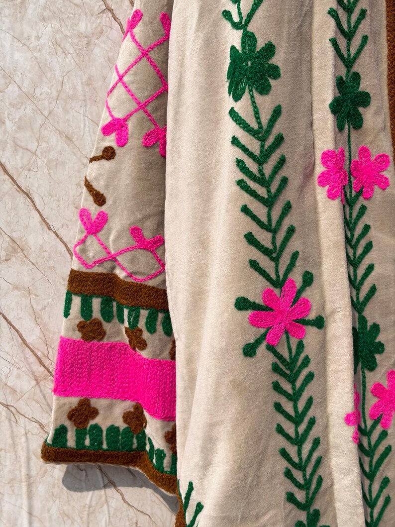 Puede incluir: Primer plano de una prenda beige bordada con detalles vibrantes. El dise&ntilde;o presenta patrones florales rosas y verdes y acentos geom&eacute;tricos. La manga tiene un patr&oacute;n de rayas marrones y rosas. La prenda est&aacute; hecha de una tela suave y texturizada.