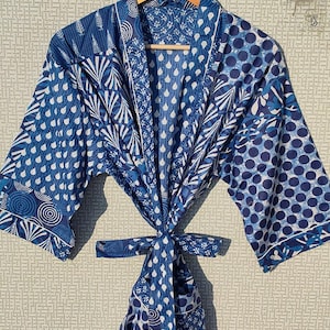 May include: A blue and white patterned robe with a matching tie, hanging on a wooden hanger. The robe features various geometric and floral designs in shades of blue and white. The robe is made of a lightweight fabric.