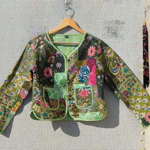 Op de afbeelding: Een patchwork jasje met een V-hals en lange mouwen. De jas heeft bloemen- en paisley patronen in groen, roze en blauw. Het heeft twee voorzakken en is gevoerd met een groen-wit gestreepte stof.