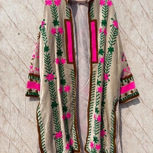 Puede incluir: Chaqueta beige sin cierre, adornada con bordados florales rosas y verdes. La chaqueta presenta ribetes marrones y un forro a rayas. Las mangas y el cuello tambi&eacute;n est&aacute;n decorados con detalles bordados.