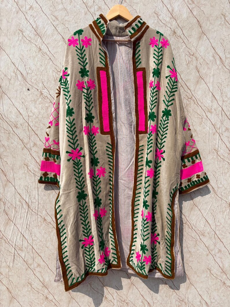 Puede incluir: Chaqueta beige sin cierre, adornada con bordados florales rosas y verdes. La chaqueta presenta ribetes marrones y un forro a rayas. Las mangas y el cuello tambi&eacute;n est&aacute;n decorados con detalles bordados.
