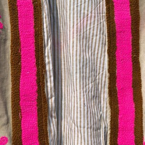 Puede incluir: Primer plano de un textil con fondo beige. El textil presenta un borde marr&oacute;n con un dise&ntilde;o rectangular rosa brillante. El interior del textil tiene un patr&oacute;n de rayas en beige y blanco.