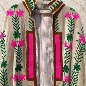 Puede incluir: Chaqueta beige bordada con cuello y ribete marr&oacute;n. La chaqueta presenta bordados florales rosa brillante y hojas verdes. El forro interior es un estampado de rayas. La chaqueta est&aacute; colgada en una percha de madera.