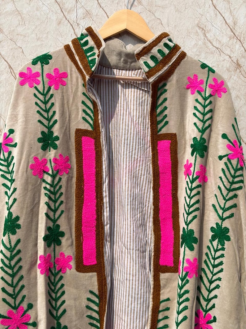 Puede incluir: Chaqueta beige bordada con cuello y ribete marr&oacute;n. La chaqueta presenta bordados florales rosa brillante y hojas verdes. El forro interior es un estampado de rayas. La chaqueta est&aacute; colgada en una percha de madera.