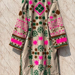 Puede incluir: Abrigo largo de terciopelo beige con intrincados bordados. El abrigo presenta patrones florales y geom&eacute;tricos rosas, verdes y marrones. Las mangas y el cuello est&aacute;n detallados con dise&ntilde;os similares, creando una est&eacute;tica bohemia.