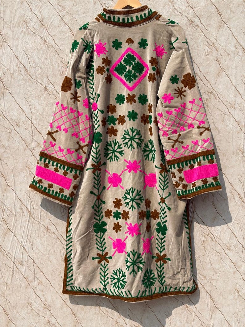 Puede incluir: Abrigo largo de terciopelo beige con intrincados bordados. El abrigo presenta patrones florales y geom&eacute;tricos rosas, verdes y marrones. Las mangas y el cuello est&aacute;n detallados con dise&ntilde;os similares, creando una est&eacute;tica bohemia.