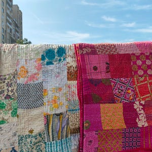 Könnte beinhalten: Zwei Patchwork-Quilts, einer in Creme und der andere in Rosa- und Rottönen, hängen vor blauem Himmel. Die Quilts bestehen aus verschiedenen Stoffmustern und -texturen und zeigen eine handwerkliche Ästhetik. Die Quilts werden im Freien ausgestellt.