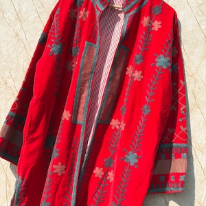 Puede incluir: Un abrigo rojo vibrante con bordados florales y de hojas en verde azulado y marrón. El abrigo presenta un forro a rayas y se muestra en una percha de madera. Las mangas largas y el diseño frontal abierto sugieren un estilo bohemio o vintage.