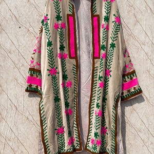 Puede incluir: Un abrigo largo y abierto de color beige neutro, adornado con bordados vibrantes. El dise&ntilde;o presenta flores rosas, hojas verdes y ribetes marrones. El abrigo est&aacute; colgado de una percha de madera, mostrando sus intrincados detalles y su silueta fluida.