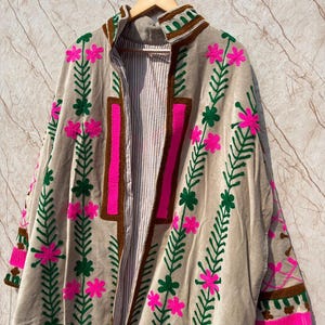 Puede incluir: Chaqueta beige de frente abierto con bordados florales. La chaqueta presenta dise&ntilde;os florales rosas y verdes, ribetes marrones y un interior a rayas. La chaqueta est&aacute; colgada en una percha de madera.