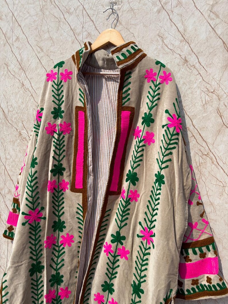 Puede incluir: Chaqueta beige de frente abierto con bordados florales. La chaqueta presenta dise&ntilde;os florales rosas y verdes, ribetes marrones y un interior a rayas. La chaqueta est&aacute; colgada en una percha de madera.