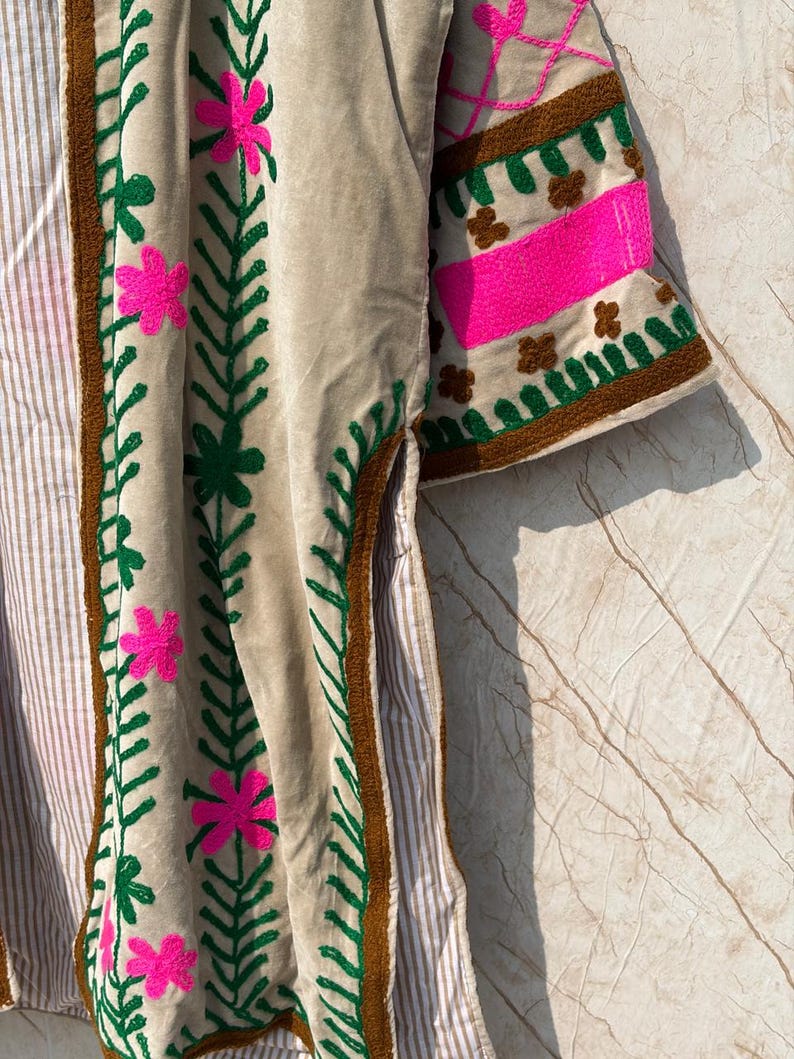 Puede incluir: Chaqueta de terciopelo beige con bordados florales. La chaqueta presenta flores rosas brillantes y hojas verdes bordadas en la parte delantera y las mangas. Las mangas tienen un ribete marr&oacute;n y una franja rosa. La chaqueta tiene un forro a rayas.