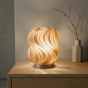 Può includere: Una lampada da tavolo decorativa con un design unico e vorticoso. La lampada è di colore beige caldo e poggia su una base metallica rotonda. Il testo "SCULPTURE + LIGHT" è visibile su una piccola etichetta. Un vaso con un ramo è accanto alla lampada.