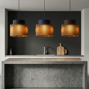 Op de afbeelding: Drie hanglampen op een rij, elk met een cilindervormige, geribbelde amberkleurige glazen kap. De bovenste delen van de armaturen zijn massief, uitgevoerd in matzwart, donkerblauw en donkerbruin, opgehangen aan dunne zwarte snoeren.