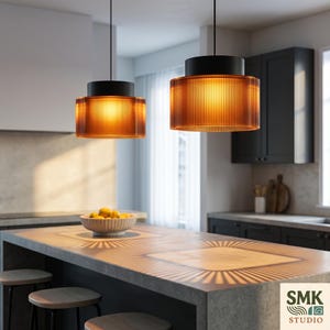 Könnte beinhalten: Zwei zylindrische Pendelleuchten mit schwarzen Oberteilen und gerippten, bernsteinfarbenen Glasschirmen. Die Leuchten strahlen ein warmes Licht aus und werfen markante, strahlenförmige Lichtmuster auf die Oberfläche der Kücheninsel.