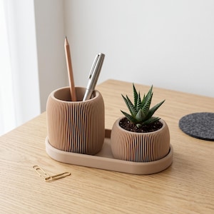 Può includere: Un set organizzatore da scrivania beige composto da un portapenne cilindrico a coste e un vaso a coste abbinato contenente una piccola pianta succulenta verde, entrambi su un vassoio ovale. Una matita in legno e una penna argentata sono all'interno.