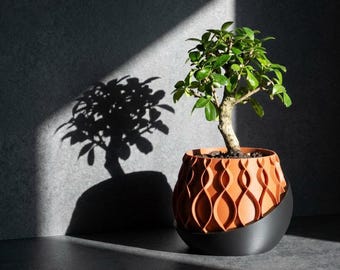 May include: A small green bonsai tree planted in a terracotta-colored pot with a repeating vertical wave pattern. The pot is nested inside a smooth, matte black curved base that cradles the lower portion of the textured orange planter.