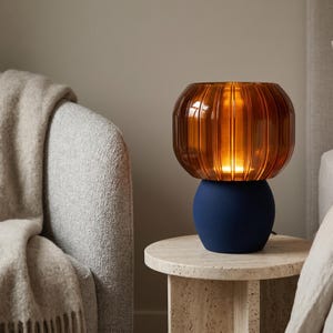 May include: A table lamp featuring a rounded, matte navy blue base and a translucent, amber-colored glass shade with vertical ribbed detailing. The lamp emits a warm, soft glow from within the glass shade, creating a distinct contrast between the dark base.