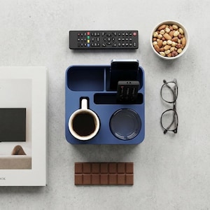 Könnte beinhalten: Ein dunkelblauer, rechteckiger Schreibtisch-Organizer mit mehreren Fächern, der eine weiße Keramiktasse mit schwarzem Kaffee, ein Smartphone, eine kleine Fernbedienung und einen runden Untersetzer enthält. Der Organizer ist umgeben von einem Buch, einer Fernbedienung, einer Schale mit Nüssen und einer Schokoladentafel.