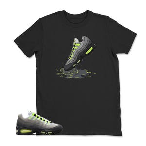 Może przedstawiać: Czarny t-shirt z grafiką szarego i neonowo-zielonego buta sportowego. But sportowy jest przedstawiony w dynamicznej pozie, z efektem rozprysku w kolorze neonowej zieleni i szarości. Pasujący but sportowy jest pokazany na dole obrazu.