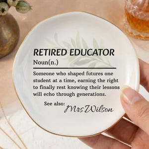 May include: A white ceramic ring dish with gold trim, featuring the text "RETIRED EDUCATOR" and a definition. The dish is held by a hand, with a gold ring and necklace visible. A small perfume bottle and a vase of flowers are in the background.