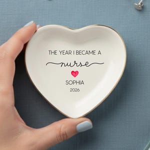 May include: Heart-shaped ceramic trinket dish with gold trim, inscribed with "THE YEAR I BECAME A nurse" and personalized with a name and year. The dish is held against a blue background, with jewelry and a small bottle nearby.