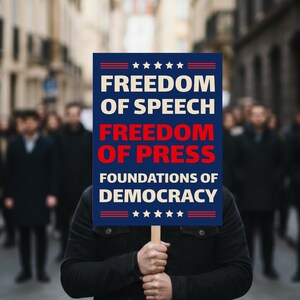 May include: A rectangular sign held by a person, featuring a dark blue background with white and red text that reads: FREEDOM OF SPEECH, FREEDOM OF PRESS, FOUNDATIONS OF DEMOCRACY, accented with white stars at the top and bottom of the sign design.