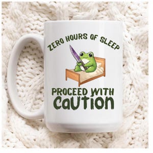 May include: White ceramic mug with a humorous design. The mug features a green frog holding a knife while sitting on a bed, with the text "ZERO HOURS OF SLEEP" and "PROCEED WITH CAUTION" in green font.