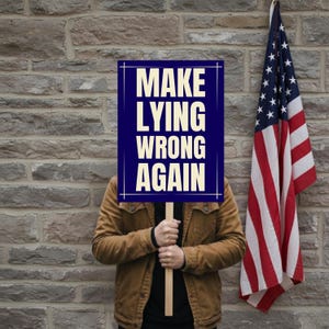 May include: A rectangular sign with a dark blue background and cream-colored text that reads MAKE LYING WRONG AGAIN. The sign is held by a person wearing a brown jacket, positioned next to a hanging American flag against a gray stone wall.