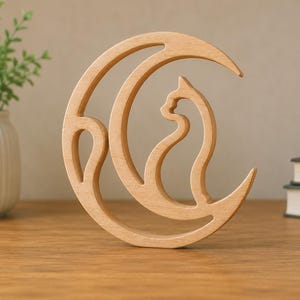 May include: A light-colored wooden cutout sculpture featuring a crescent moon shape with a cat silhouette integrated into the center. The smooth, natural wood grain is visible across the entire surface of this decorative object, which stands upright.