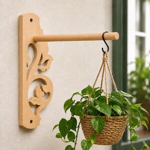 May include: A light-colored wooden wall-mounted plant hanger featuring a decorative cutout scrollwork bracket and a horizontal rod. A black metal S-hook suspends a woven jute basket containing a green leafy plant from the wooden rod.