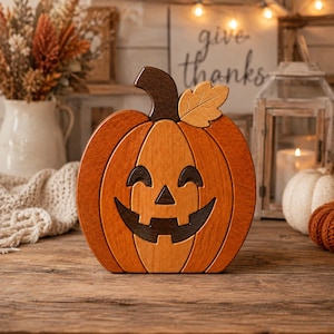 May include: A wooden jack-o'-lantern decoration featuring a carved pumpkin shape with a dark brown stem and a single light brown leaf. The pumpkin has a smiling face with dark brown eyes, a triangular nose, and a wide, jagged mouth.