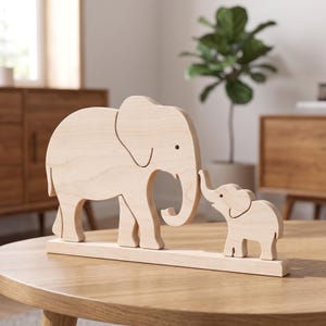 Op de afbeelding: Een lichtgekleurd houten beeldje met een grote olifant die naast een kleine olifant staat. Beide figuren zijn bevestigd op een rechthoekige houten voet en hebben een gladde afwerking met een natuurlijke houtnerf en strakke contouren.