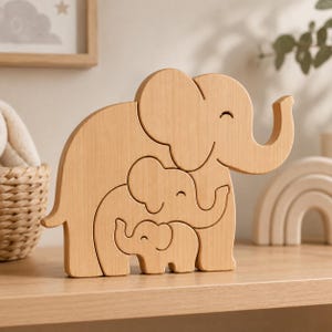 以下が含まれることがあります： 3つの入れ子状のゾウの形をした明るい色の木製パズル。一番大きなゾウのシルエットの中に中くらいのゾウがあり、その中にさらに小さなゾウの形が収まっています。それぞれのゾウには、彫り込まれたシンプルな目と口があります。