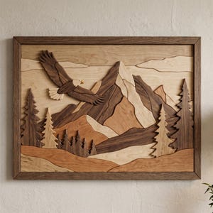 Op de afbeelding: Houten wandkunst met een vliegende adelaar boven een bergketen. Het kunstwerk is gemaakt van verschillende houttinten, waaronder donkerbruin, lichtbruin en beige, waardoor een gelaagd effect ontstaat. Het stuk is ingelijst in donker hout.