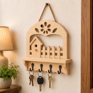 May include: A light-colored wooden wall-mounted key holder featuring a cutout flower design at the top, a small birdhouse and picket fence motif, and a shelf. Four black metal hooks are attached to the bottom, each holding a metal key ring.