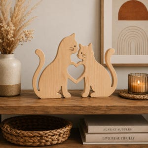 May include: A light-coloured wooden decorative sculpture featuring two stylised cats facing each other. Their front paws meet to form a heart-shaped cutout in the centre. The cats have smooth, clean lines and a natural wood grain finish, standing upright.