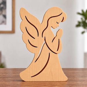 May include: A light-coloured wooden cutout of an angel in a kneeling position with hands pressed together in prayer. The figure features smooth, curved lines defining the wings, flowing gown, and head, showcasing a natural wood grain texture.