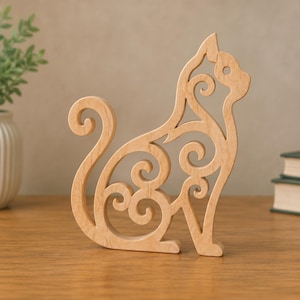 Könnte beinhalten: Eine hellfarbene Katzenfigur aus Holz mit einem komplizierten durchbrochenen Design, das durchgehend wirbelnde, schnörkelartige Muster an Körper, Schwanz und Kopf aufweist. Die Katze ist im sitzenden Profil mit geringeltem Schwanz und glatten Kanten dargestellt.